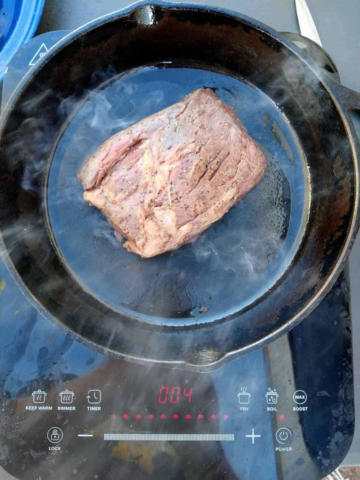 A cast iron skillet on a induction cooktop with a Bavette steak being seared in it. You can see smoke rising from the skillet.