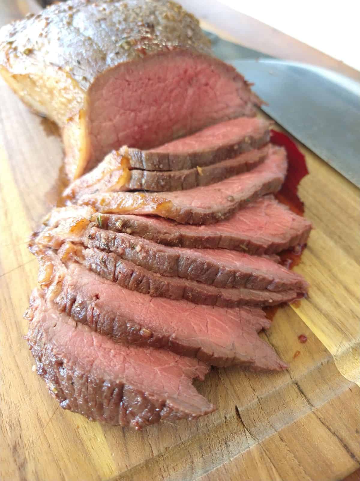 Slices of browed bottom round roast that is pinkish red on the inside are on top of a wood cutting board. A knife is to the right of the roast.