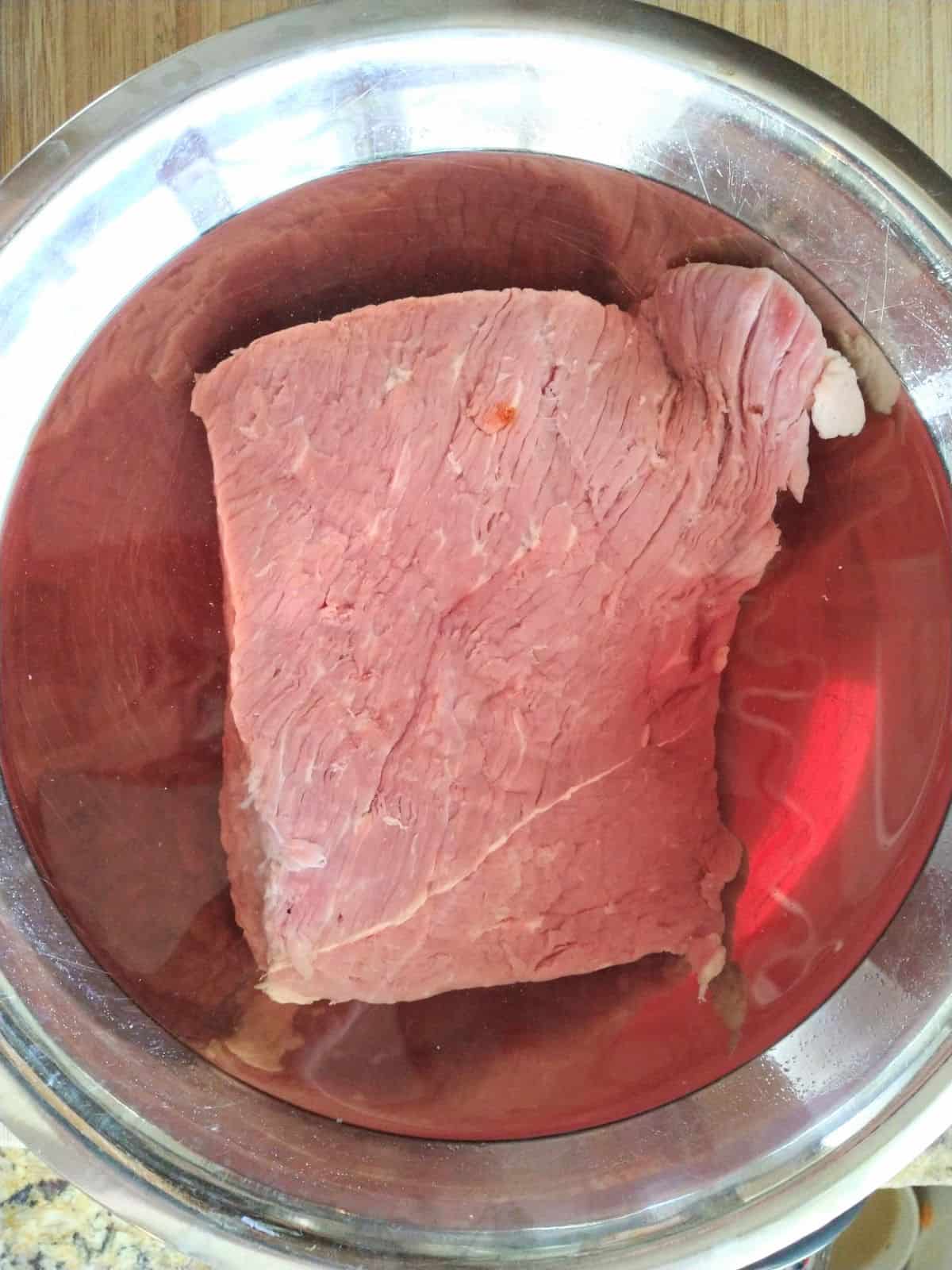 Corned beef that has been placed in a mixing bowl filled with water for 3 hours. The water has a pink hue to it. 