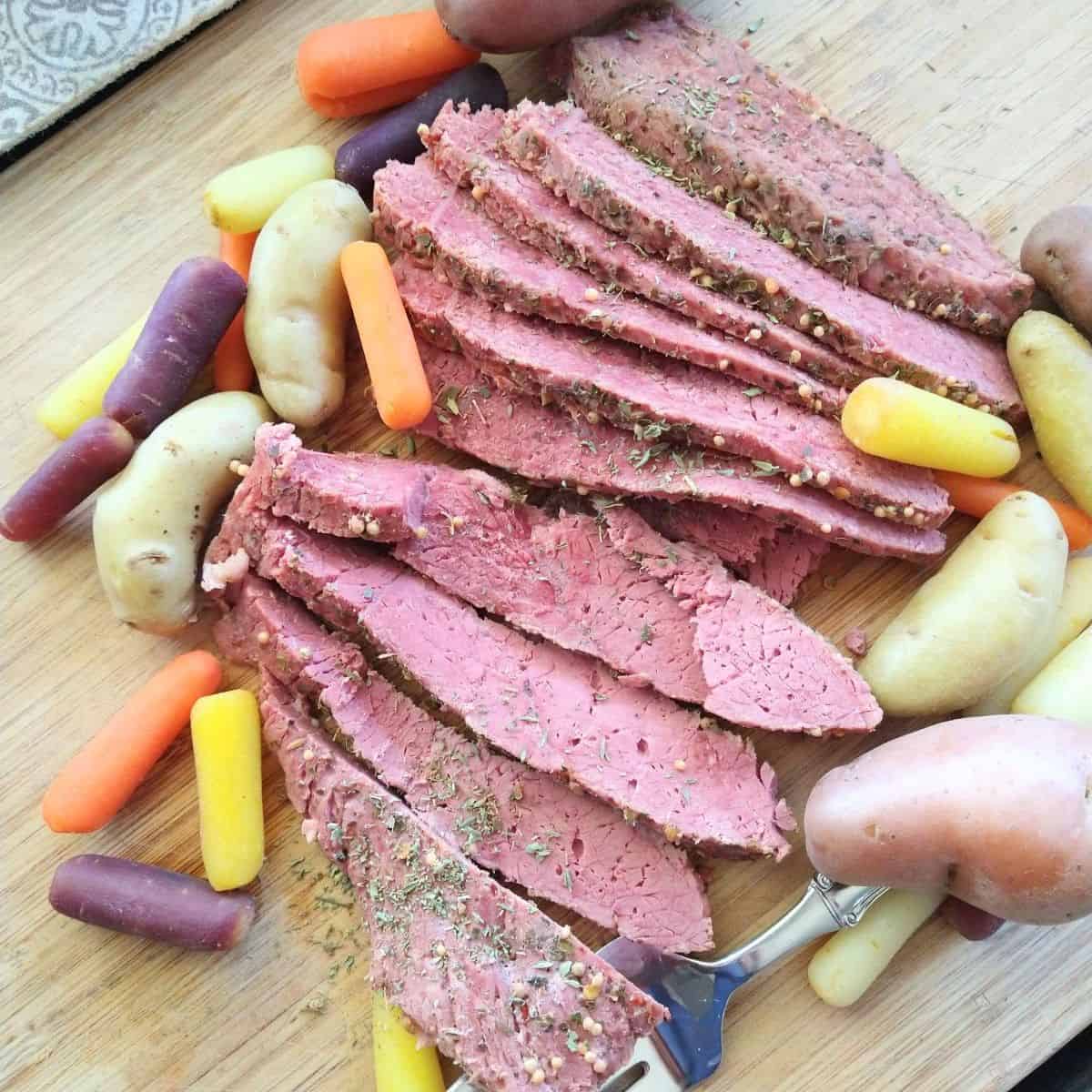 Cooked Corned beef arranged in slices on a wood cutting board with fingerling potatoes and rainbow baby carrots. 