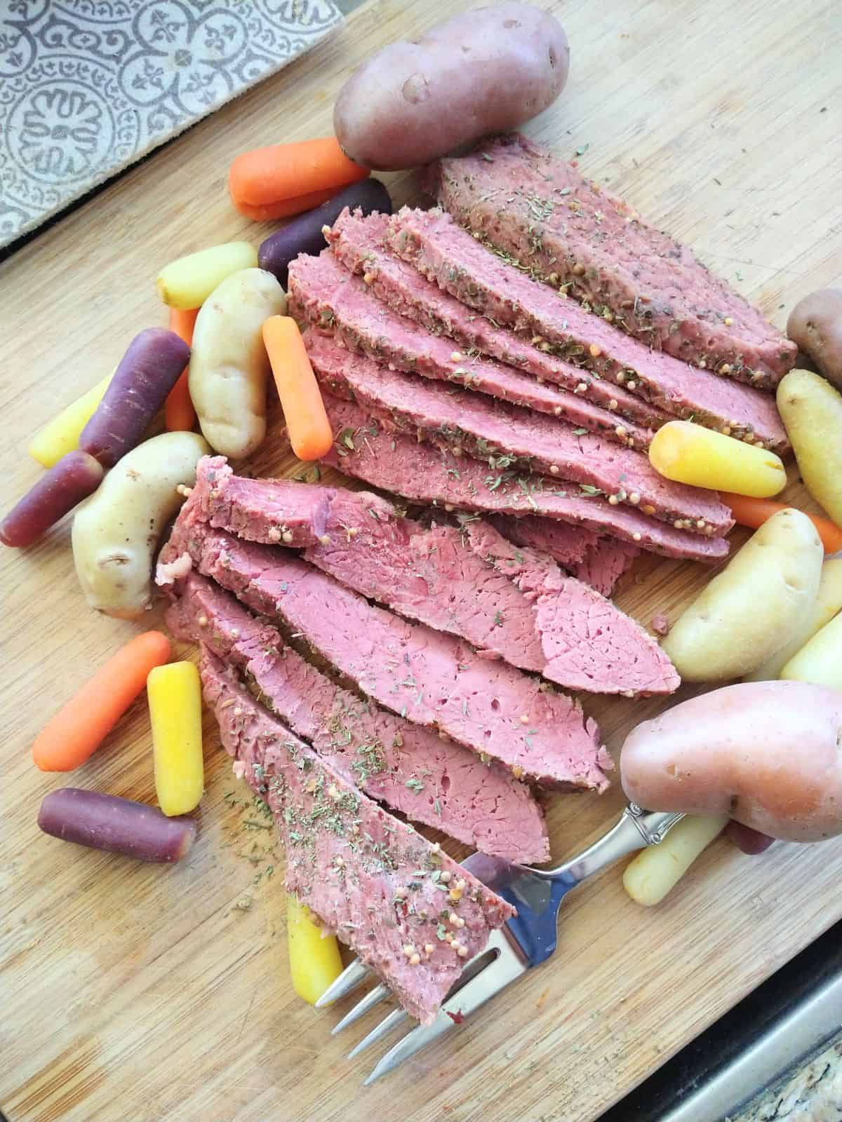 Slices of cooked corned beef with spices on the outside are stacked up on a cutting board with rainbow baby carrots and white and red fingerling potatoes.