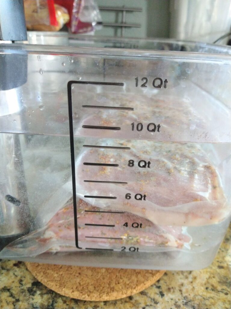 Two packages of vacuum sealed Corned beef on top of each other inside plastic container water bath with a sous vide machine attached. You can see qt measurements on the side of the container.