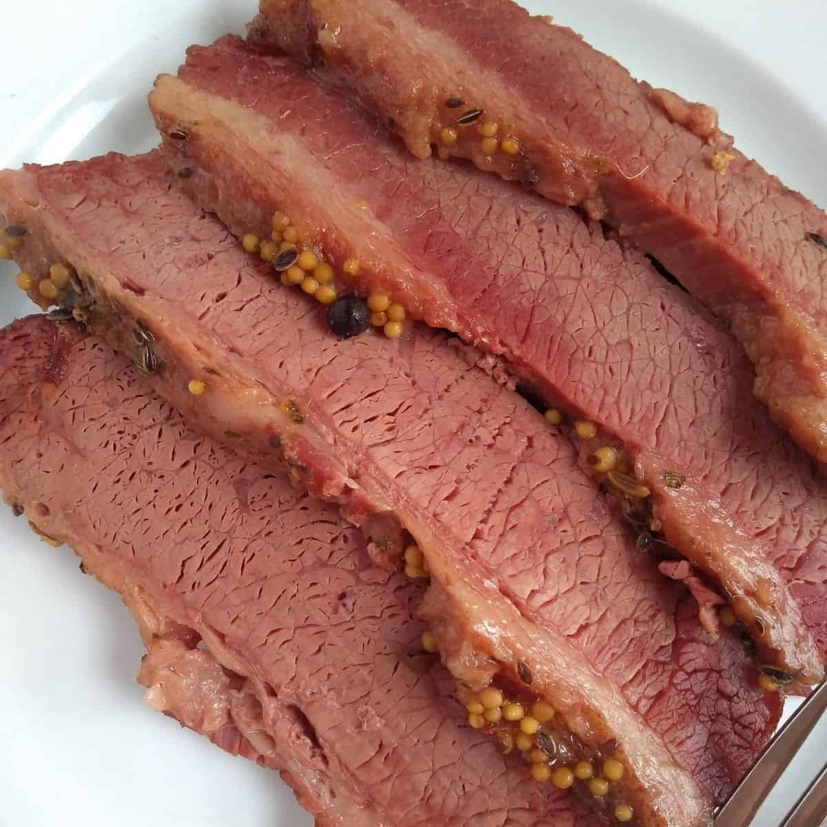 A close up of corned beef brisket slices on a white plate. You can see the whole pickling spices on the side of the meat.