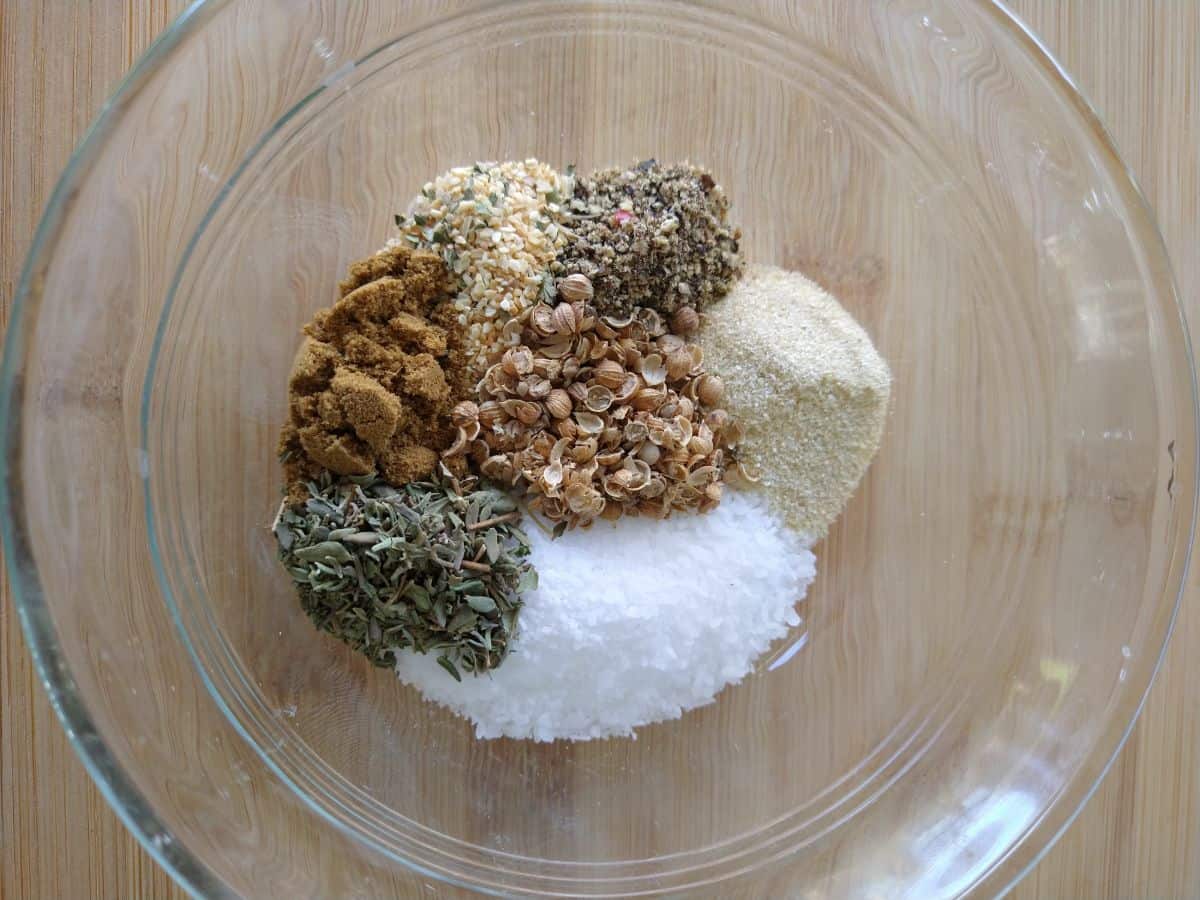 Spices and seasonings are pilled separately in a glass bowl. The spices include garlic powder, kosher salt, dried thyme, crushed coriander, ground cumin, and freshly cracked peppercorns.