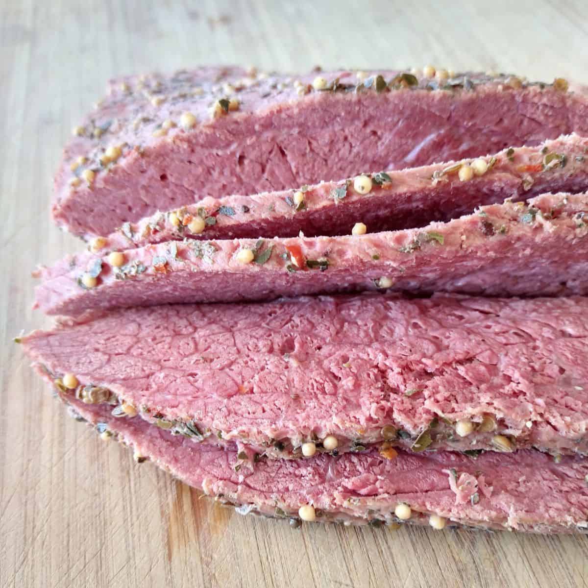 Sliced corned beef with spices on the outside laying on a wood cutting board.