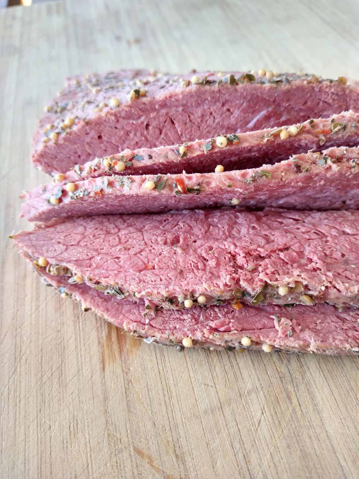 Slices of corned beef sitting on a wood cutting board. You can the spices on the edge of the meat.