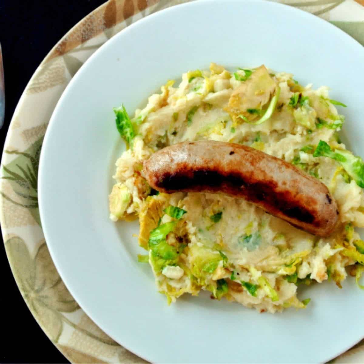 Mashed potatoes with Brussel sprouts mixed in with a Irish banger sausage on top all sitting on a white plate on top of a leaf textured plate.