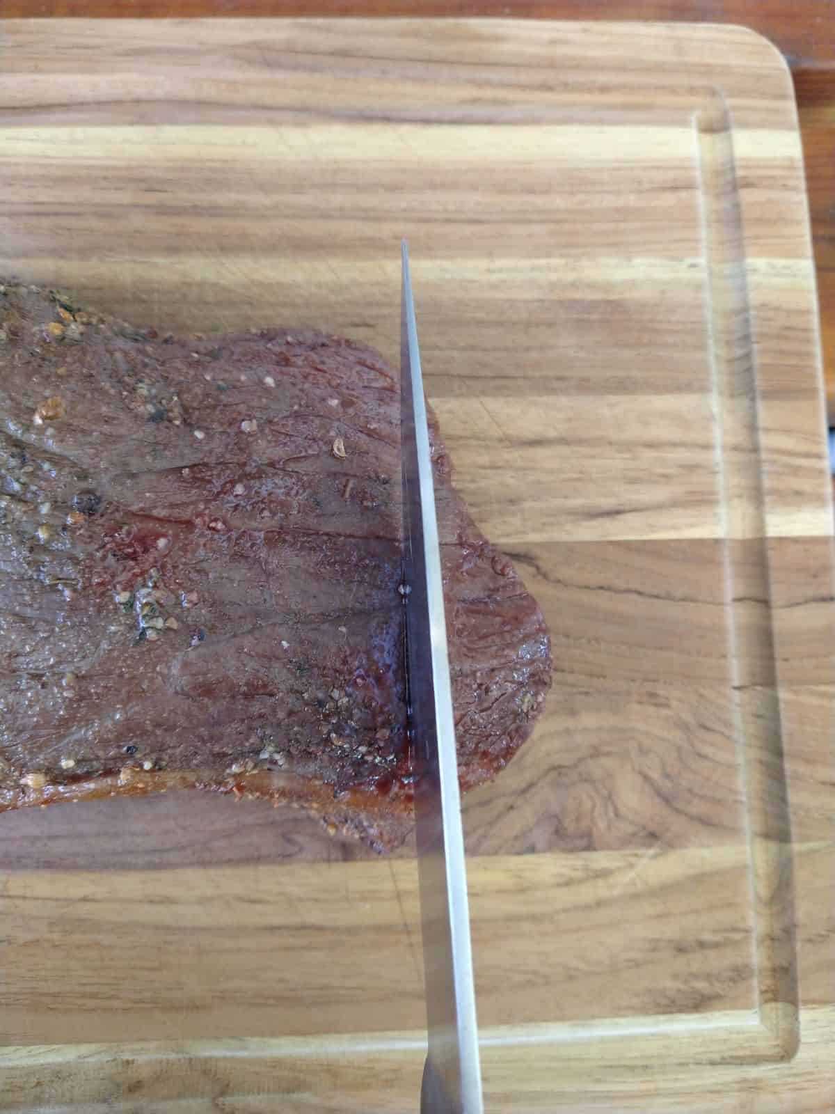 The roast is sitting on a wood cutting board with a knife on top of it that is running against the grain of the meat.