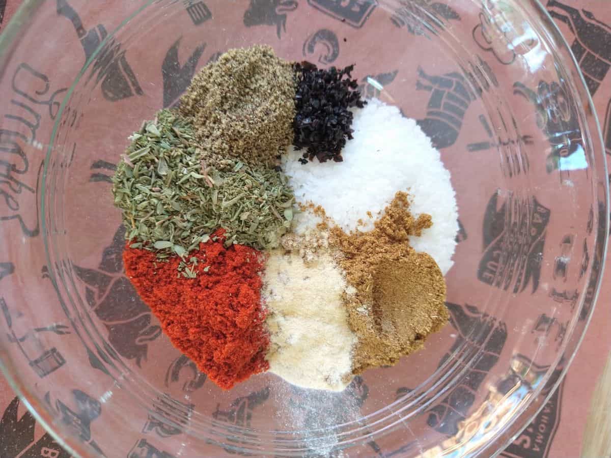 A glass bowl sitting on butcher paper filled with different spices - black urfa chili, smoked paprika, garlic powder, ground cumin, dried thyme, kosher salt, freshly ground black pepper.