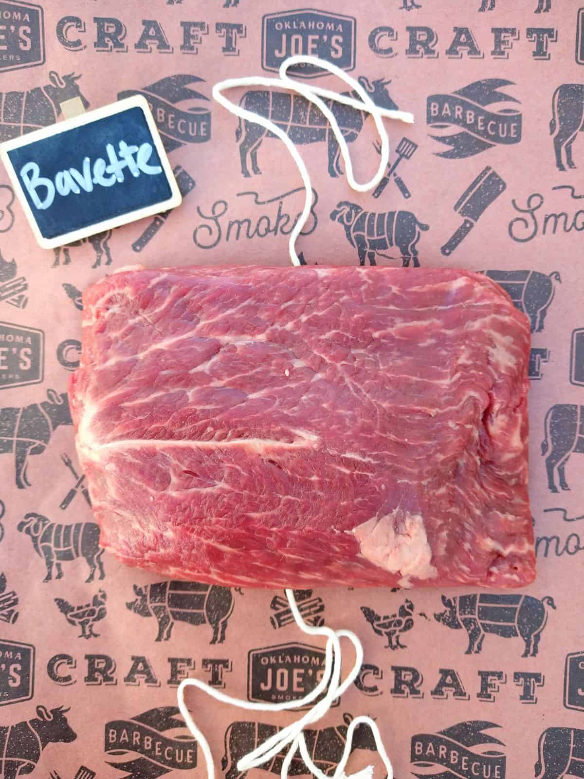 A raw Bavette steak on a maroon color piece of butcher paper with string underneath the steak and a small chalkboard sign that says "Bavette".