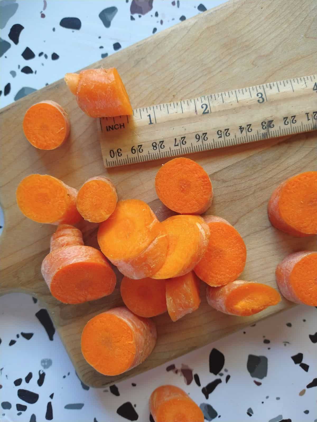 Sliced raw carrots on a cutting board with a ruler showing how think the slices are. They are about ½ to ¾ inch thick.