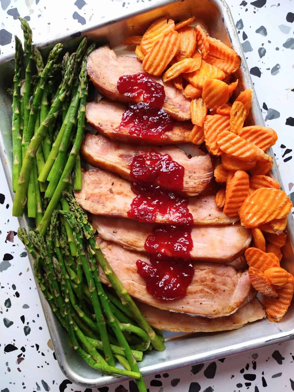 Cooked sliced ham on a sheet pan with asparagus and crinkled cut carrots. The ham has tart cherry ham on it. The sheet pan is sitting on a white table. 