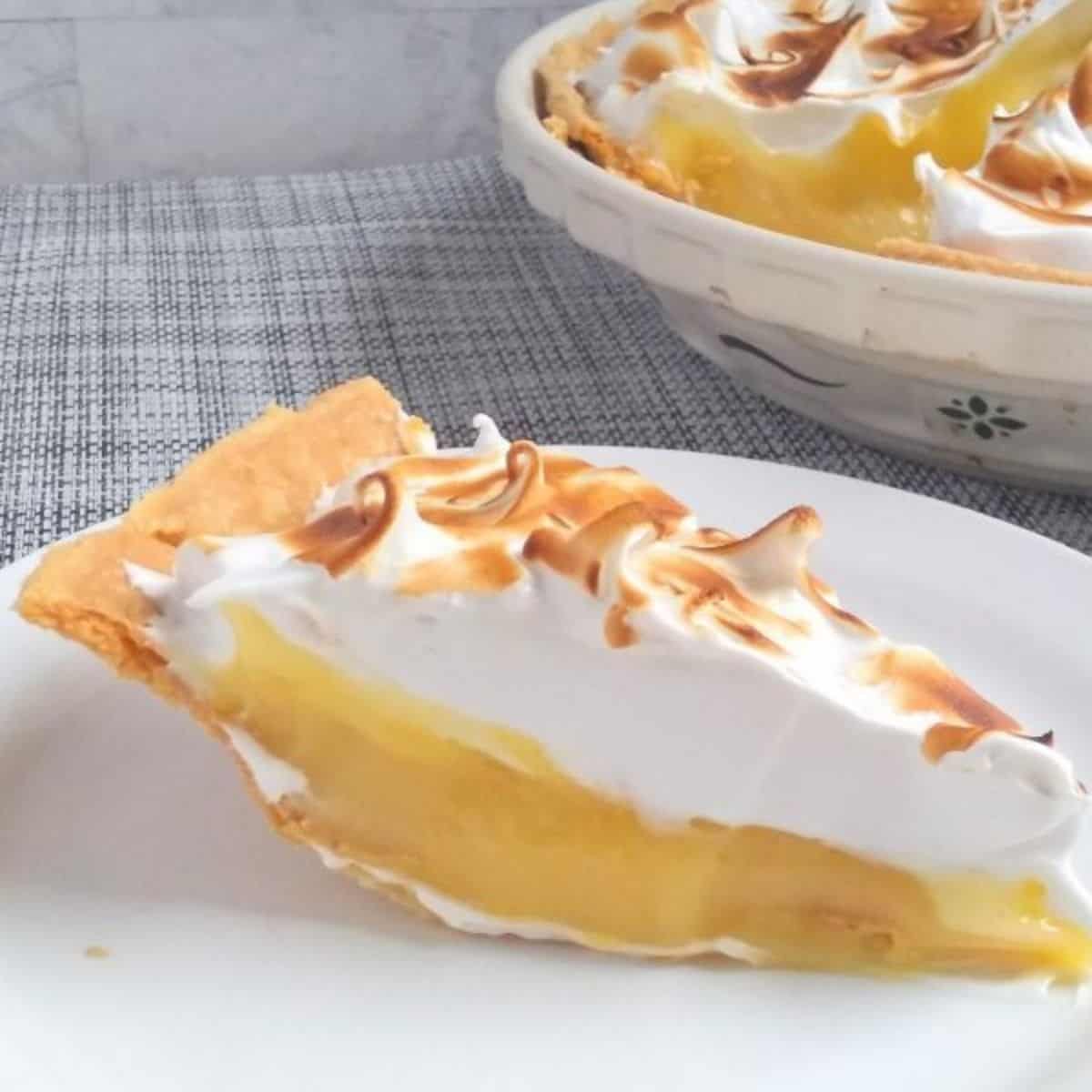 A slice of Lemon Meringue pie that has been torched on top sitting on a white plate with the whole pie in the background.