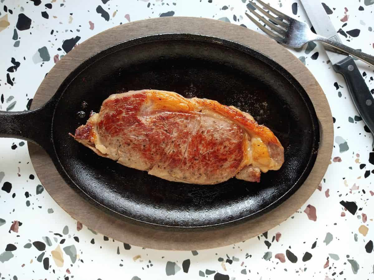 A cooked New york strip steak on a oval shaped cast iron skillet on top of a oval shaped piece of wood with a knife and fork at the top right corner.
