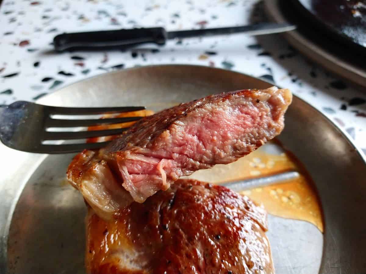 A fork holding up a piece of strip steak showing that it's pink on the inside. The rest of the steak is below on a metal plate.