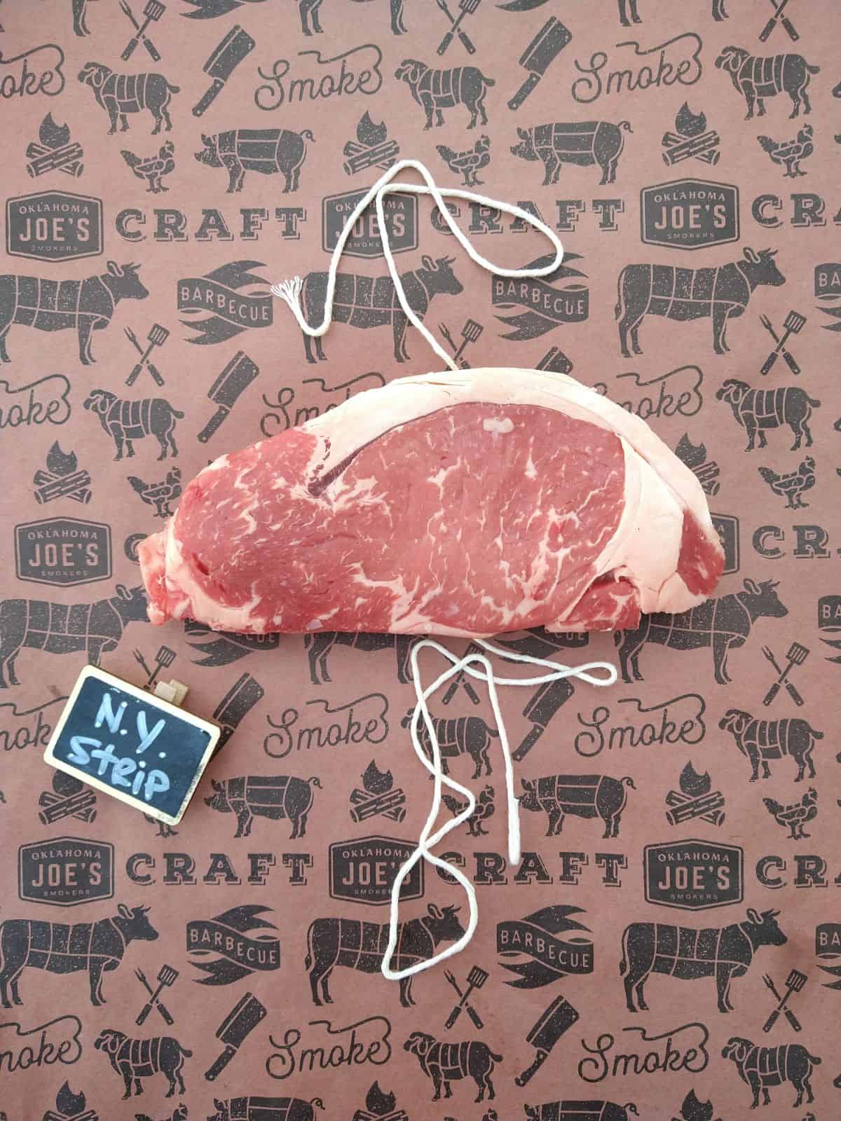 A raw NY Strip steak with a layer of fat on top, sitting on a piece of butcher paper and string. A small sign next to the steak says N.Y. Strip.