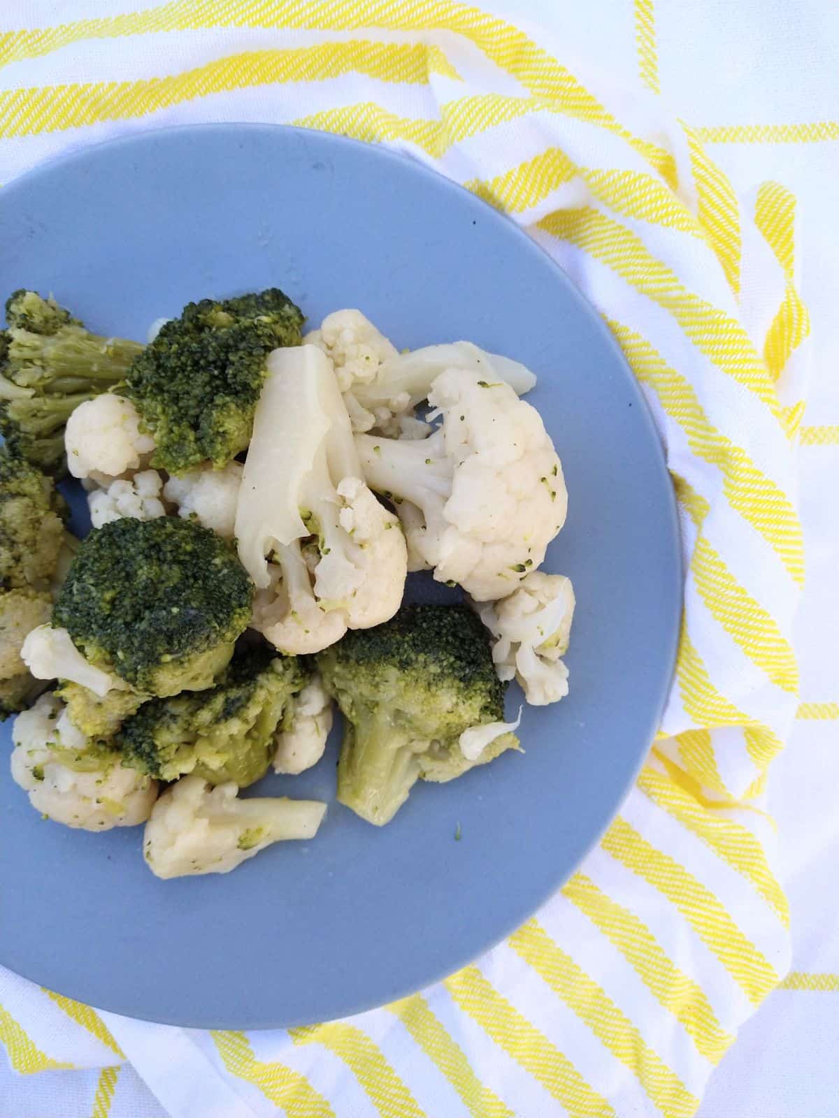 Sous vide cooked broccoli and cauliflower on a blue plate on top of yellow and white striped towels. 