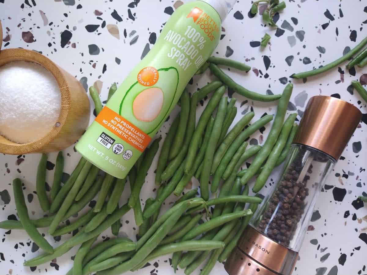 On a table sits on open salt cellar with kosher salt in it, 100% avocado spray oil, raw greens beans, and a pepper grinder with black peppercorns inside.