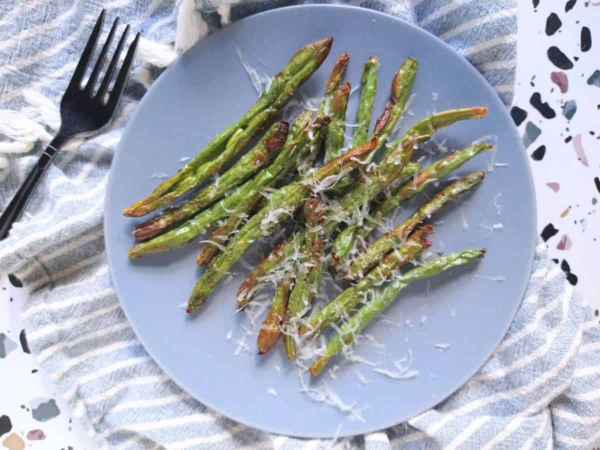 Green beans that have been air fried, on a blue plate topped with parmesan cheese. The plate is on a gray and white strip towel with a black fork next to the plate.