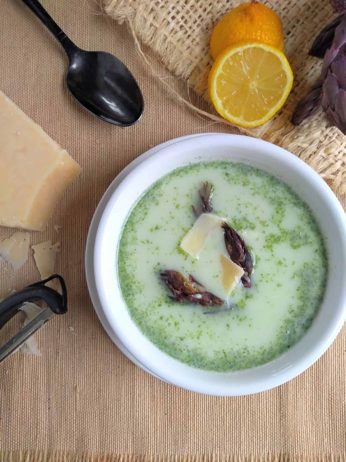 Green Asparagus ends soup topped with parmesan cheese and roasted asparagus ends on a brown placement with a black spot, half lemon, and parmesan wedge next to the bowl.