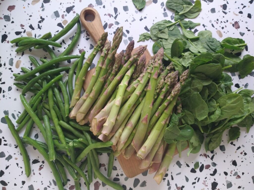 Asparagus on a small wood cutting board with green beans on the left and baby spinach on the right.
