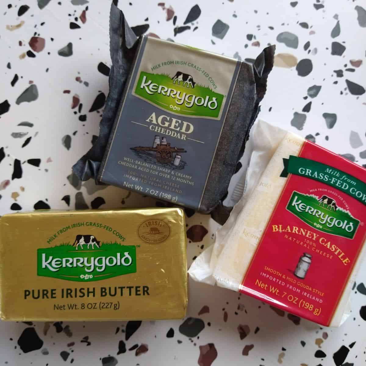 Kerrygold Products on a white table with gray and brown circles. The products are Irish butter, Aged Cheddar, and Blarney Castle cheese. 