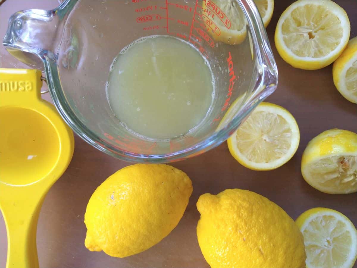 A cutting board with sliced and whole lemons, a measuring cup with lemon juice in it, and a lemon squeezer. 