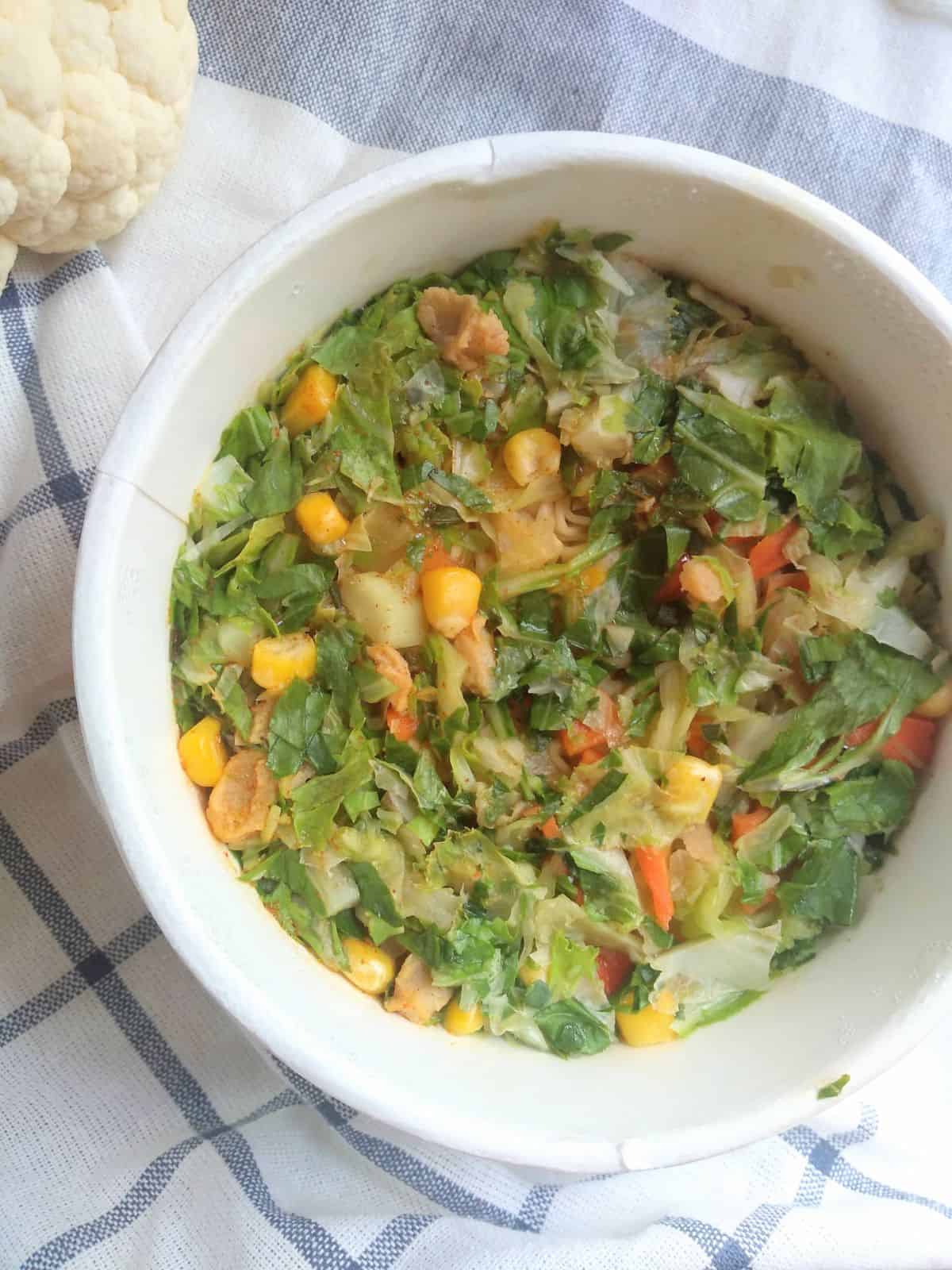 A white bowel of Ramen soup with corn, carrots, and cauliflower leaves on a white and blue striped towel.