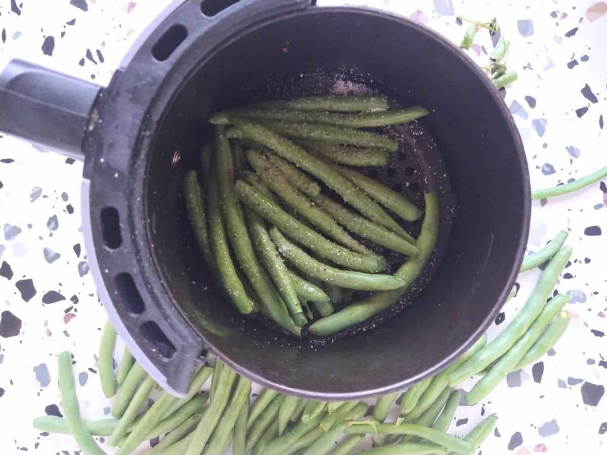 Raw green beans with salt and pepper on them inside of a basket of an air fryer.