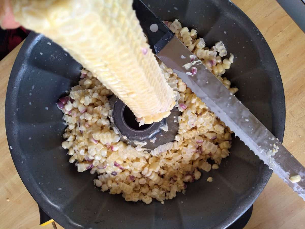 Using a bundt pan to strip corn off the cob. The corn falls into the pan. A bread knife is shown cutting the corn off. 