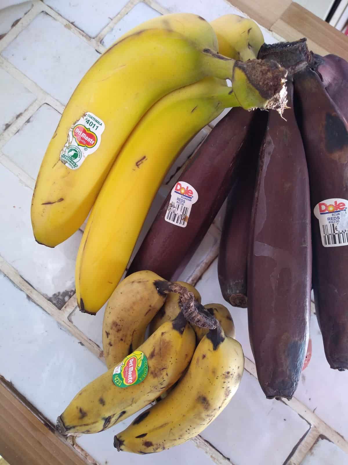 Bunches of yellow bananas, red bananas, and Manzano or apple bananas sitting on a white title countertop.