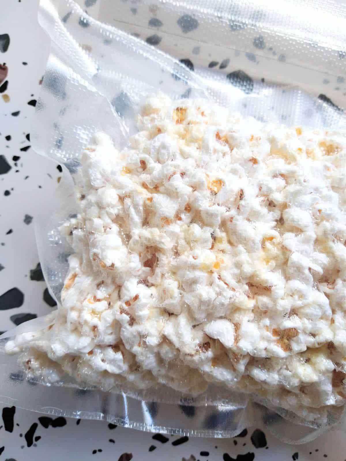 Fresh popcorn is vacuum sealed in a plastic bag, sitting on a table that is white with gray and brown spots.