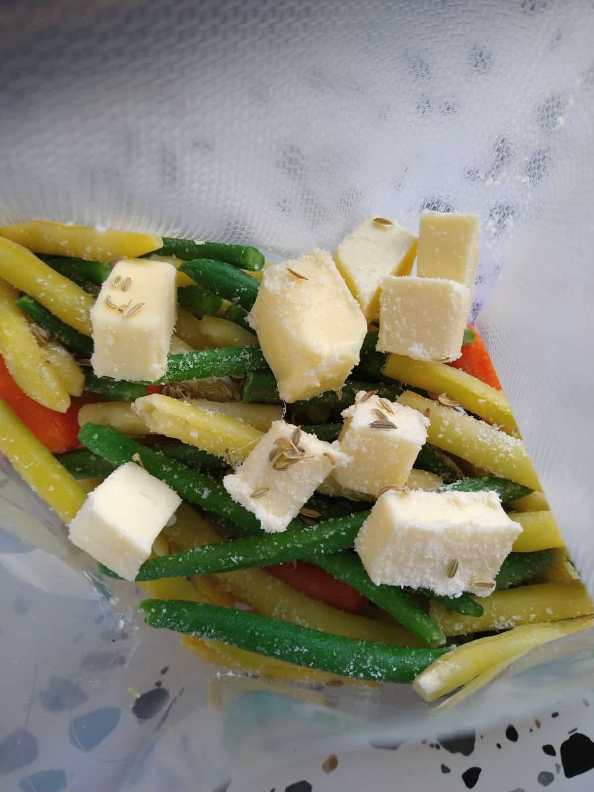 An opened plastic sous vide bag with cold butter, frozen wax beans, green beans, and baby carrots along with fennel seeds.