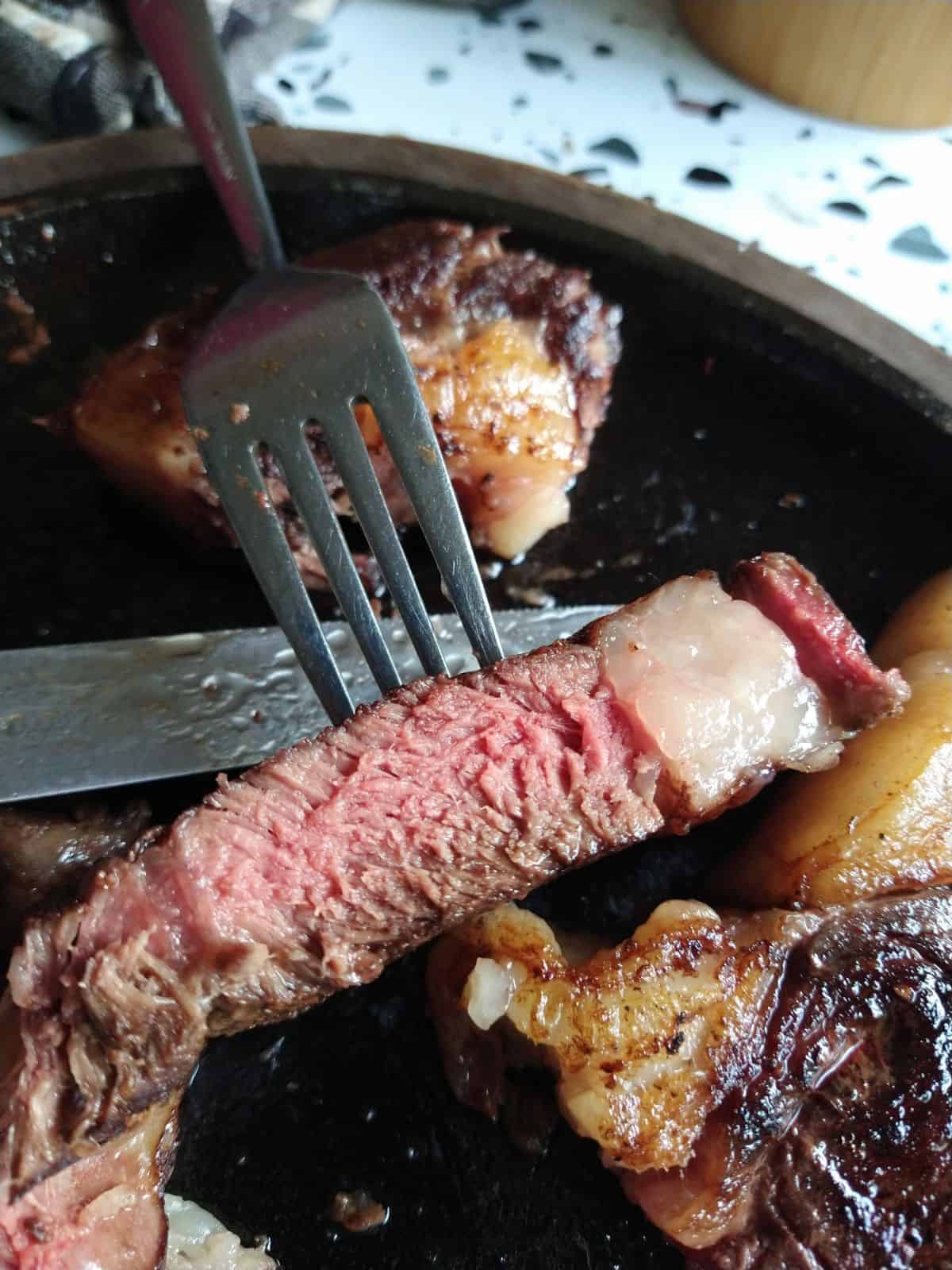 A slice of Bison ribeye cut open to show that it's pink inside. The slice is held with a fork overtop a cast iron skillet.