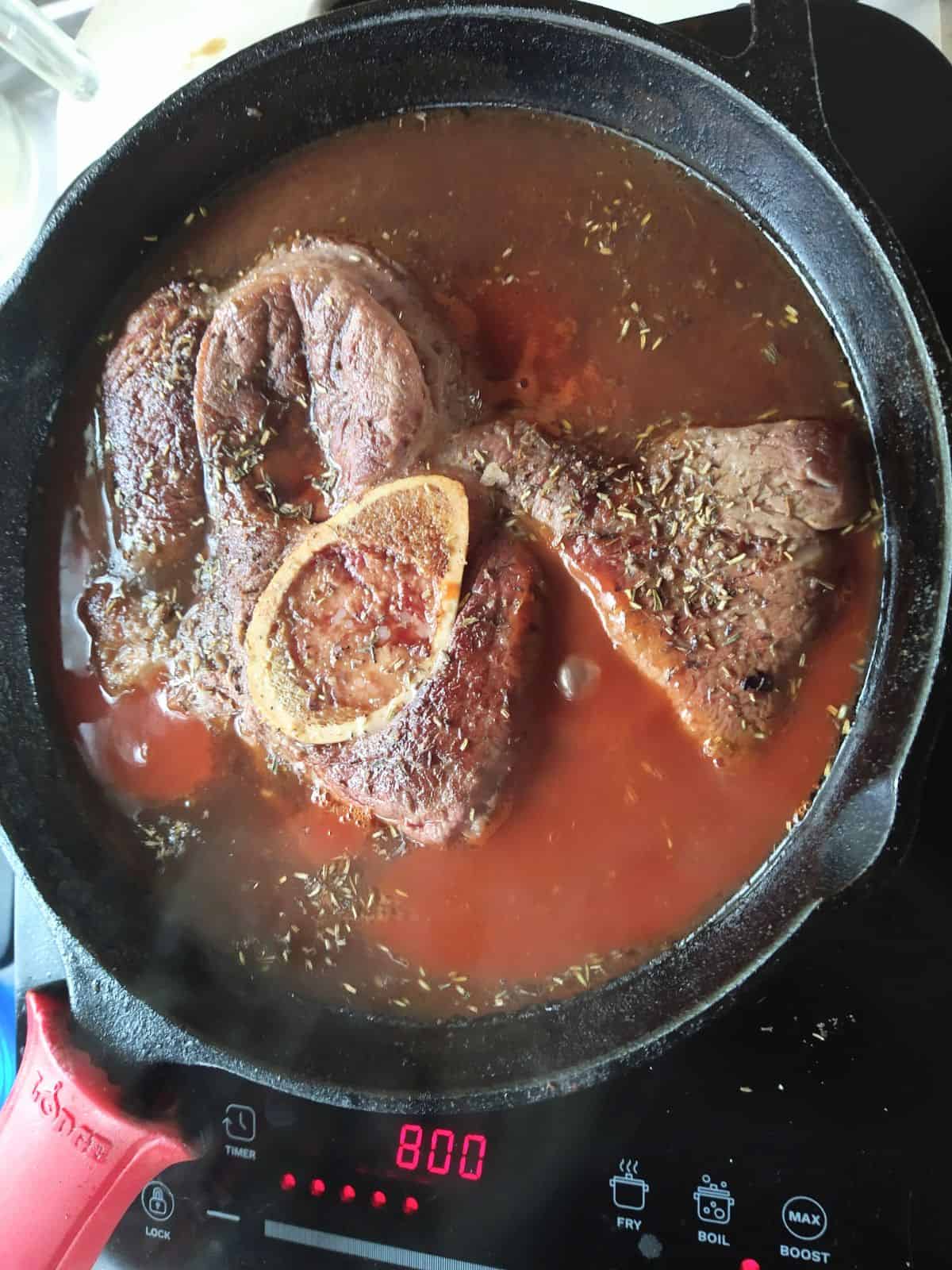 A chuck arm steak cooking in a tomato based liquid in a cast iron pan.