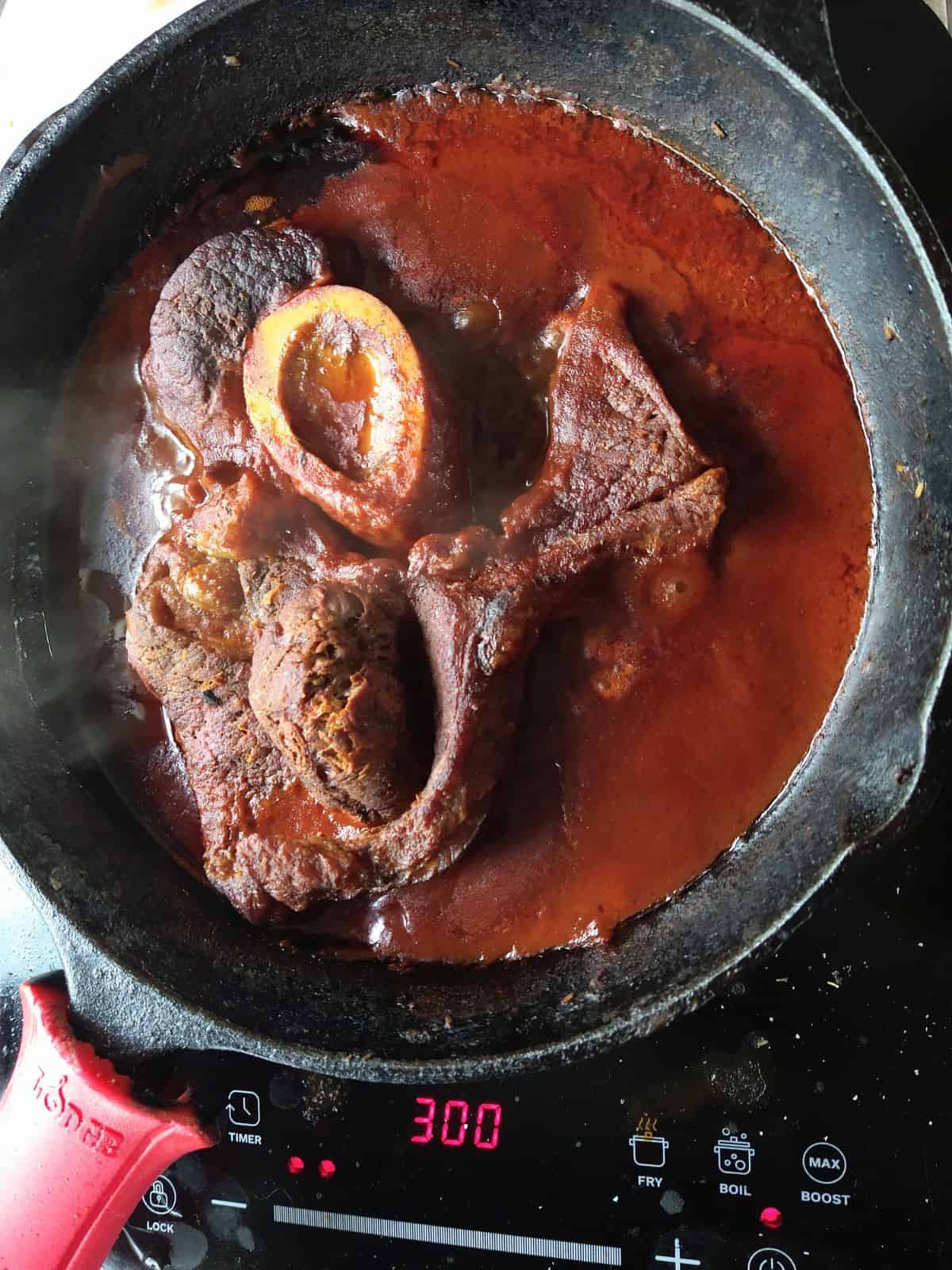 A chuck arm steak finishing cooking in a tomato based liquid in a cast iron pan.