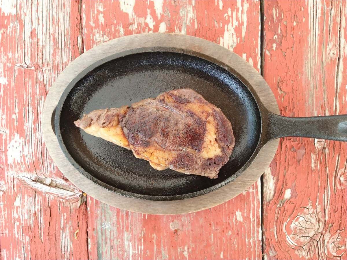 A finished and seared sous vide Bison ribeye steak on a cast iron skillet with wood holder on top of a red distressed picnic table. 