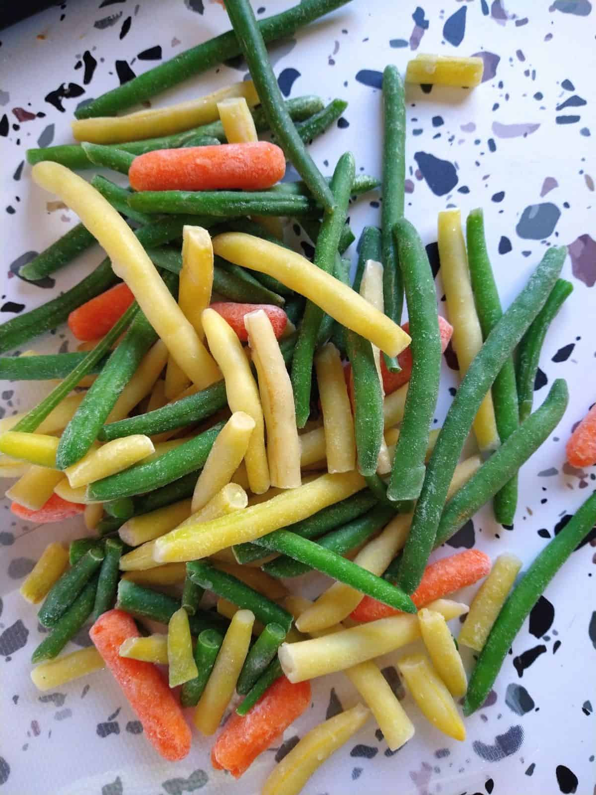 Frozen green beans, wax beans, and baby carrots sitting on a table with a white surface with colored spots.