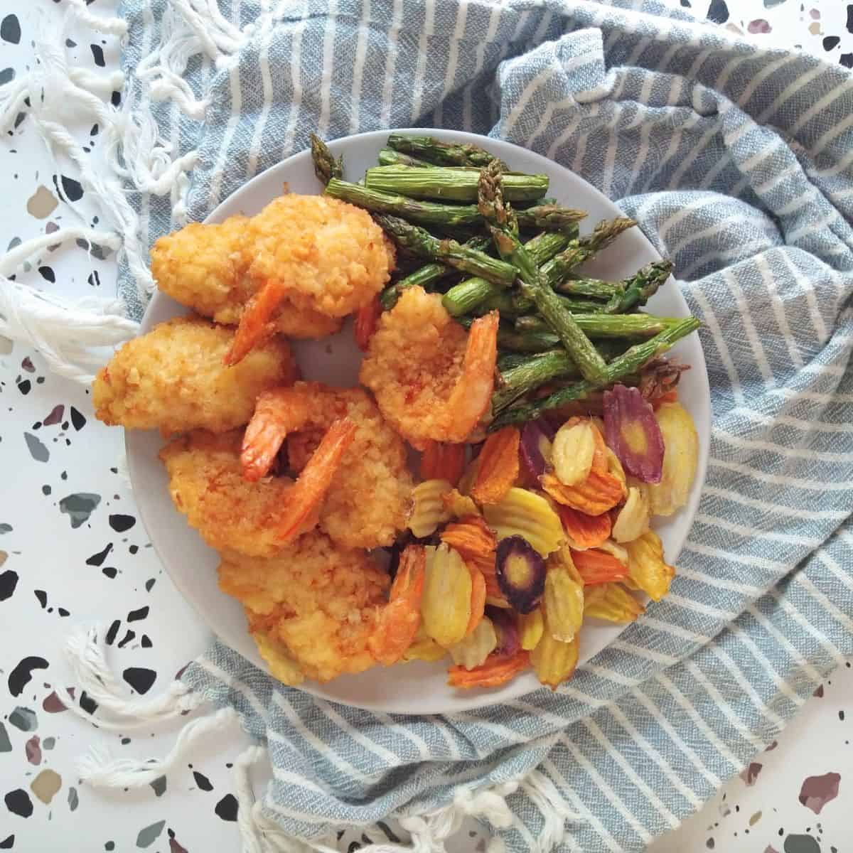 A gray plate filled with panko fried shrimp, asparagus and colorful crinkle cut carrots. The plate is on a gray striped towel that is on a white table with colored spots.
