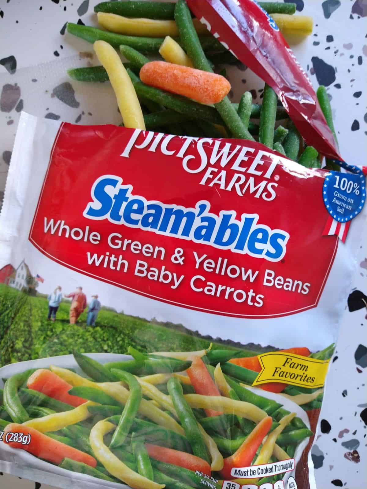 A bag of PictSweet Farms Whole Green and Yellow Beans with baby Carrots spilled out onto a white table with colored spots. 