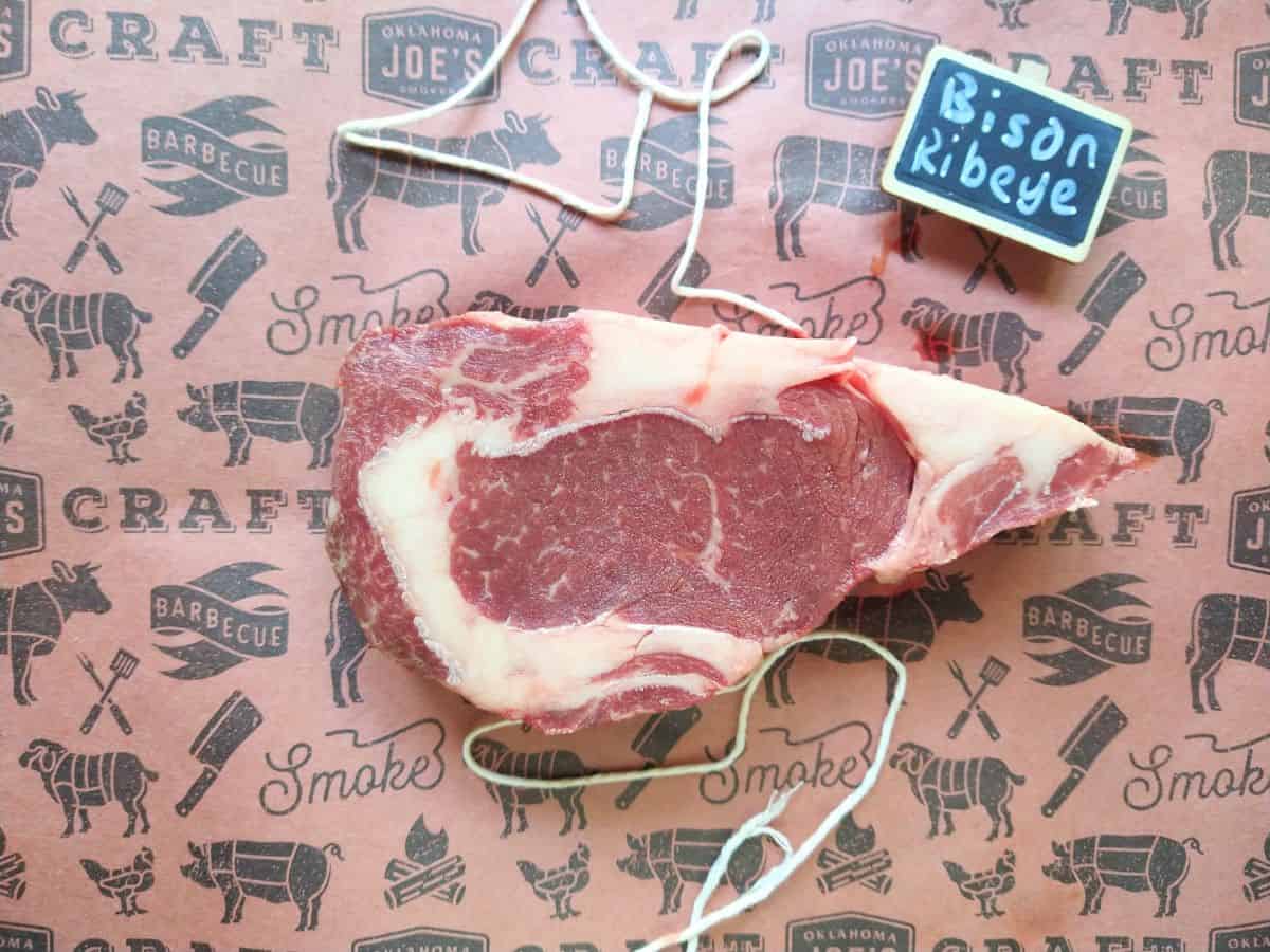 A raw Bison ribeye steak on a piece of butcher paper with a string underneath it and a small chalkboard sign that says Bison Ribeye.