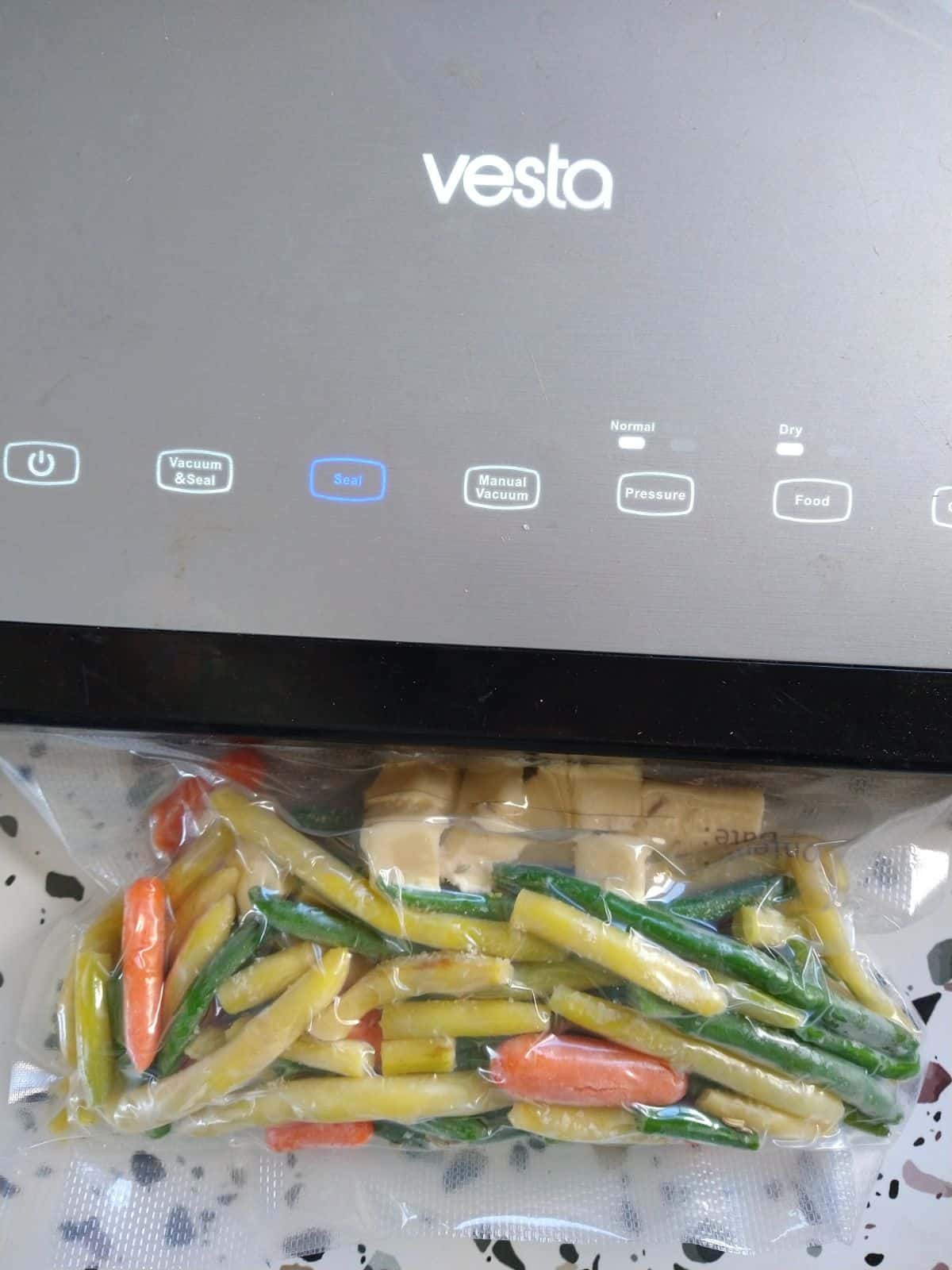 A Vesta vacuum sealer sealing a bag of baby carrots, green beans, and wax beans with butter at the top of the bag.