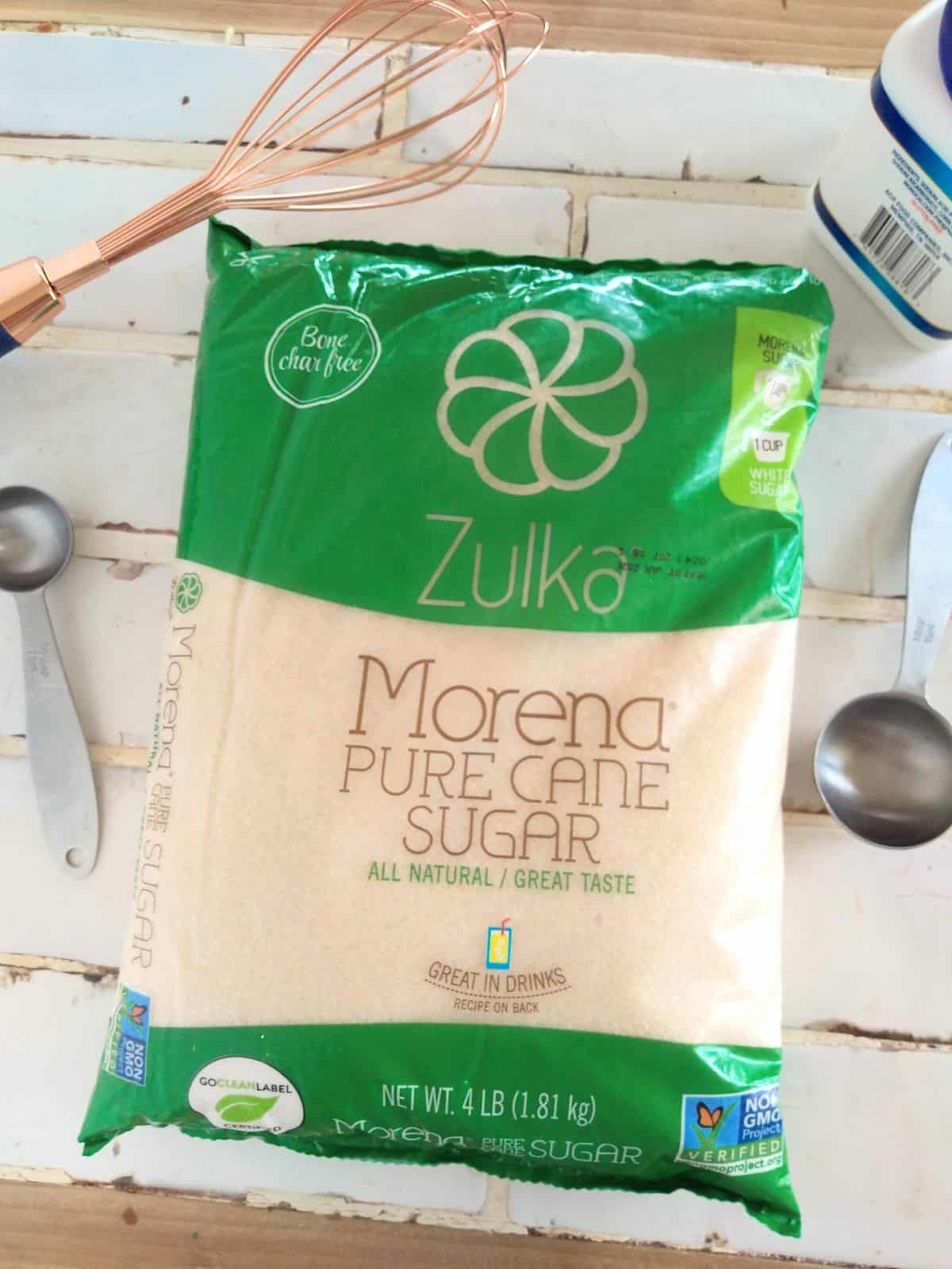 A bag of Zulka Morena Pure Sguar Bone Char Free sugar on a title countertop with measuring spoons, a whisk, and baking powder surrounding it.