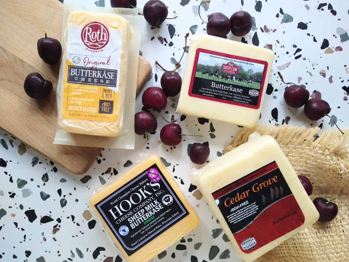 4 types of Butterkase cheese blocks (Roth, Hook's Sheep Milk, Cedar Grove, and Sassy Cow) are sitting on a white table with spots along with some fresh cherries, a small cutting board, and a piece of burlap.