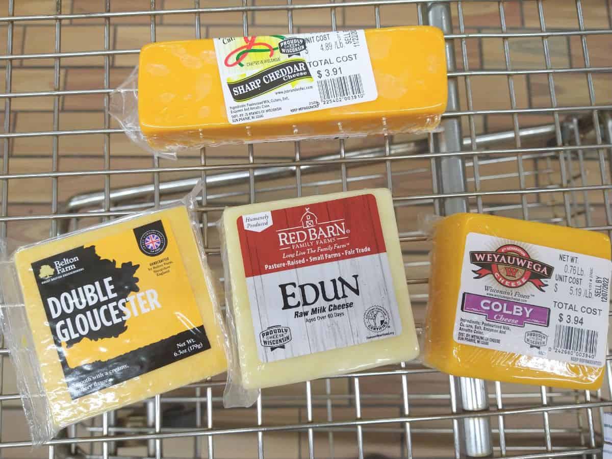 A metal grocery cart with Sharp Cheddar cheese, Double Gloucester, Edun cheese, and Colby Cheese in it.