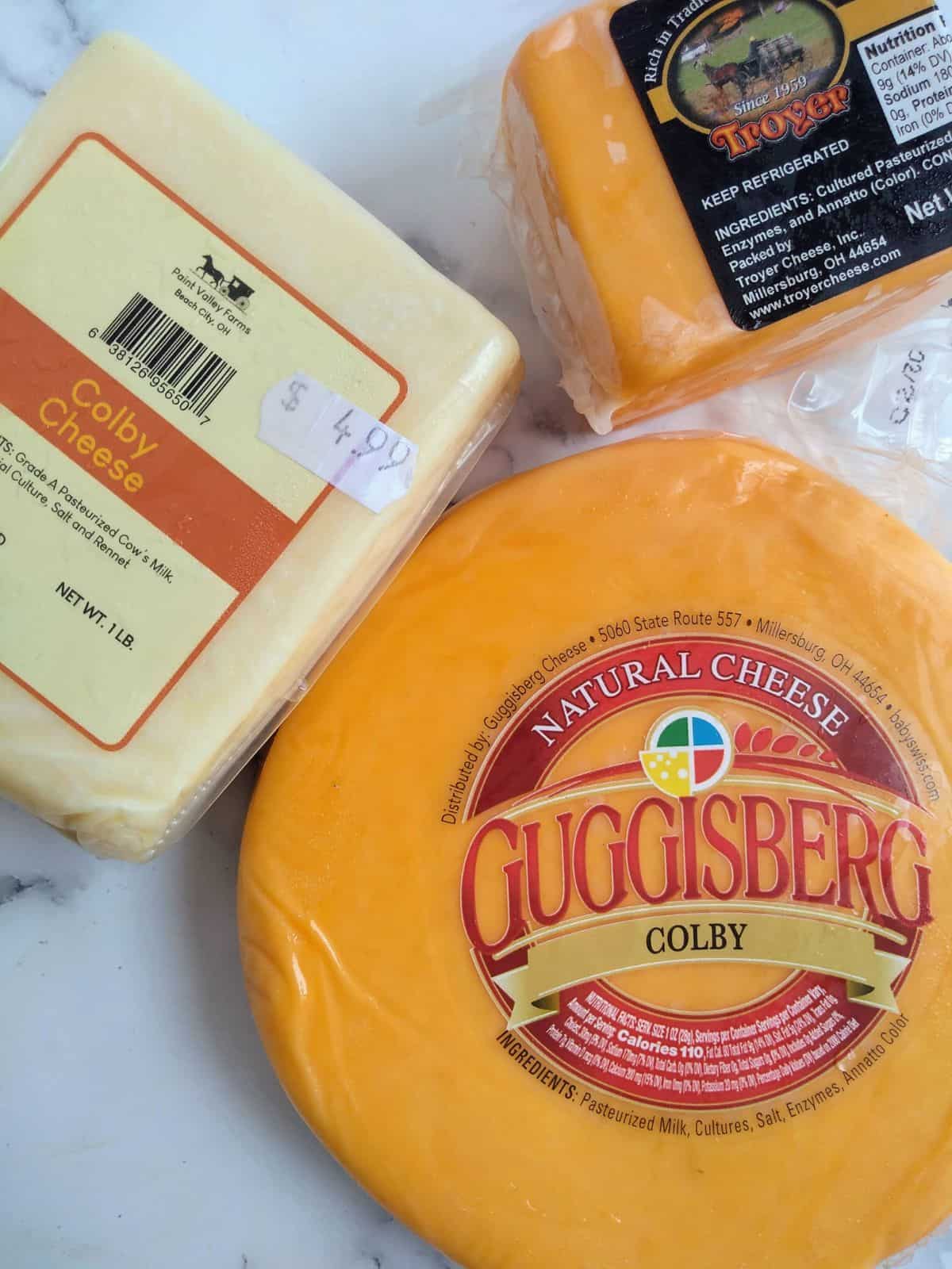 Three different brands of Colby - Pint Valley Farms, Guggisberg, and Troyer sitting on a countertop. 