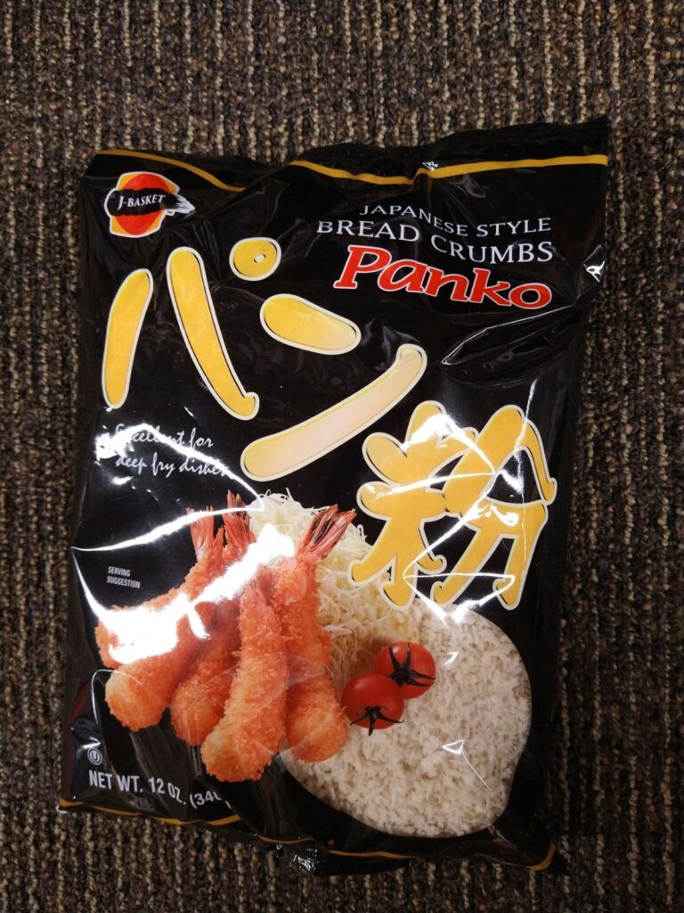 A black bag of J-Basket Japanese Style Panko breadcrumbs sitting on black carpet. The bag has a "see through window" at the bottom corner so you can see what the Panko looks like.