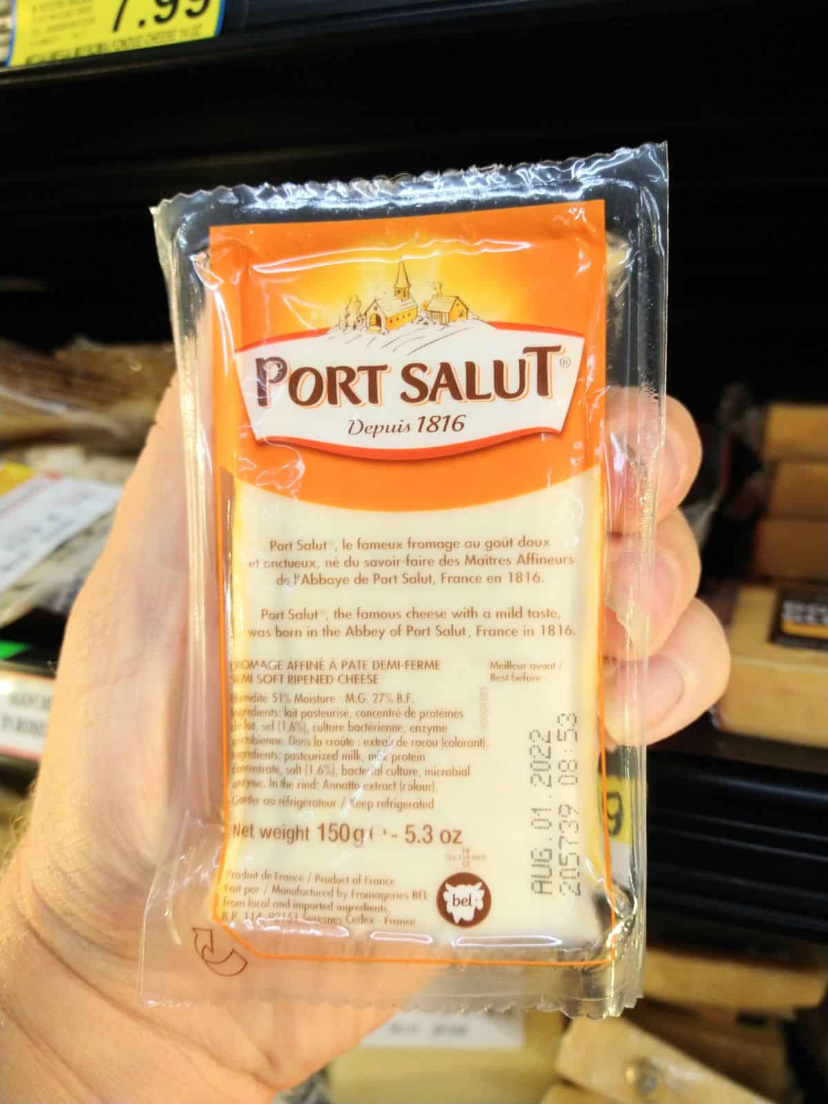 Someone holding a package of Port Salut at the grocery store in front of a display of cheese.