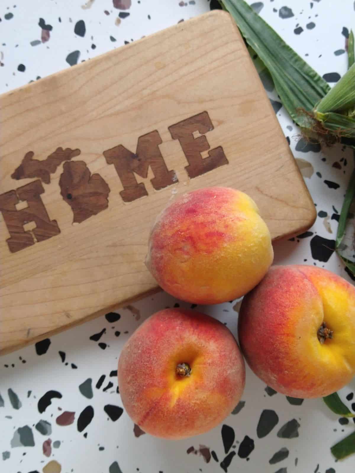 Yellow flesh peaches sitting next to a wood cutting board that says home with a picture of Michigan replacing the "O". 