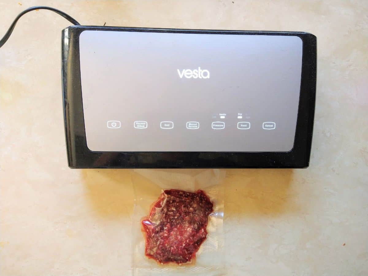 A gray and black Vesta vacuum sealer that is sealing up a single hamburger patty.